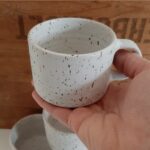 Liten rak handdrejad kopp i ljus lera med små bruna lavafläckar.
